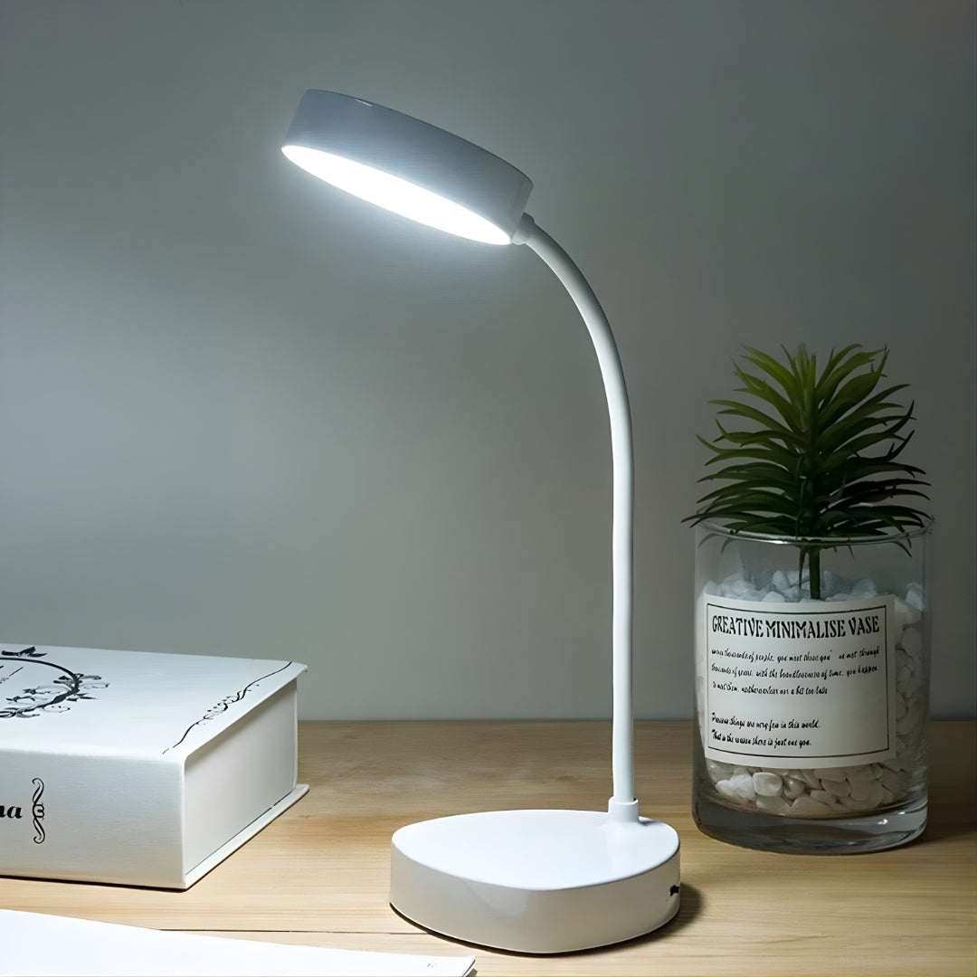 White desk lamp on a wooden surface with a book and plant in the background